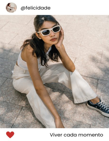 "Jovem sentada no chão, vestindo óculos, jeans e tênis pretos, em um ambiente urbano."